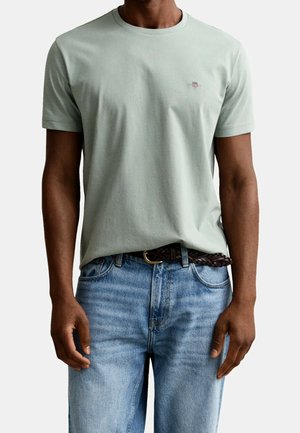 Man wearing a light green short-sleeve t-shirt tucked into light blue jeans with a brown woven belt.