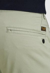 Detailný záber na béžovú kapsu nohavíc s čiernym gombíkom a štítkom PME Legend American Classic, skombinovanú s tmavomodrým vrchom.