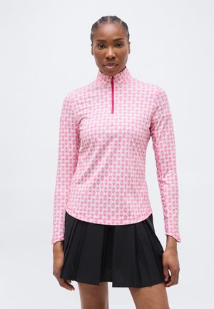 Naine, kes kannab roosa-valge mustriga pika varrukaga särki tõmblukuga krae ja musta volditud seelikut, lihtsa tausta ees.