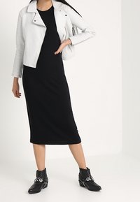 Biała skórzana kurtka motocyklowa z prążkowanymi detalami, czarna midi sukienka oraz czarne botki za kostkę z dekoracyjnymi elementami z metalu.