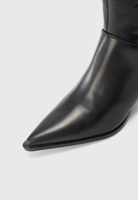 Μαύρο δερμάτινο μποτάκι αστραγάλου με μυτερή μύτη, λεία υφή, ελάχιστη ραφή και λεπτή, κομψή σχεδίαση, τοποθετημένο σε λευκό φόντο.