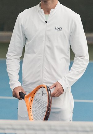 Mann in weißer Sportbekleidung hält einen orangen Tennisschläger auf einem blauen Hallentennisplatz, von der Taille aufwärts gesehen.