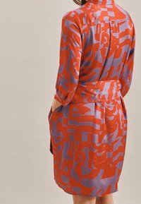 Vista trasera de una mujer que lleva un vestido de manga larga con un patrón abstracto en rojo y púrpura, con las manos en los bolsillos.