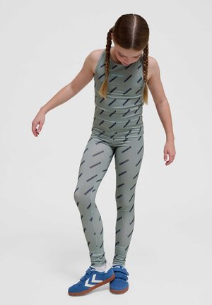 Młoda dziewczyna z zaplecionymi włosami, ubrana w szary bezrękawnik i legginsy z powtarzającym się tekstem marki, patrzy w dół, stoi na jednej nodze w niebieskich trampkach.