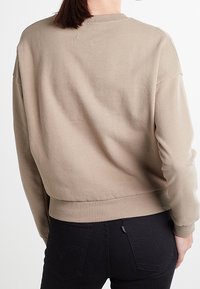 Sudadera beige de cuello redondo con un acabado texturizado, hombros caídos y puños de canalé. Lleva puesta sobre unos jeans negros de tiro alto.
