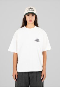 Hvit bomulls T-skjorte med korte ermer og rund hals, med en liten svart logo på brystet. Kombinert med en kremfarget caps med grønn brem.