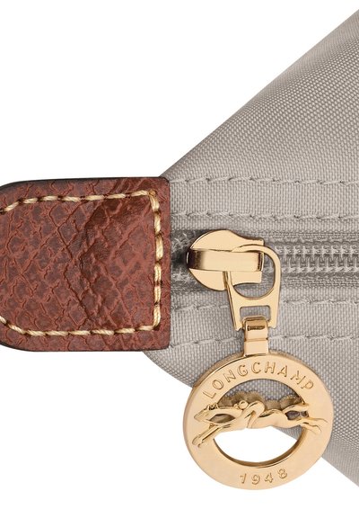 Grey fabric bag corner with textured brown leather accent, gold zipper pull with a charm featuring a running horse, and embossed branding.