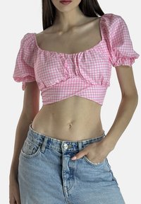 Kvinde iført pink og hvid ternet crop top med puffede ærmer og højtaljede lyseblå denimjeans, hånd i lommen.