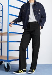 Donkerdenimjack met gouden stiksel, grijze crewneck t-shirt, zwartebroek en groene sneakers met gele strepen, staande naast een blauwe metalen rek.