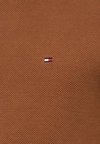 Brauner gestrickter Pullover mit einem strukturierten Muster und einem kleinen, in Blau, Weiß und Rot gestickten Logo auf der linken Seite.