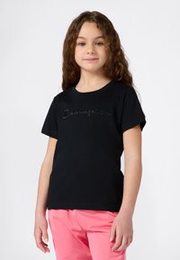 Chica con pelo rizado largo, vistiendo una camiseta negra de Champion y pantalones rosas, de pie frente a un fondo blanco liso.