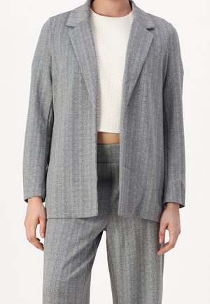 Woman wearing a gray herringbone blazer and matching pants paired with a white textured cropped top, standing against a white background.