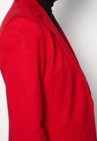 Czerwony dopasowany żakiet o gładkiej fakturze, z wyraźnie zarysowanymi ramionami i klapami. Widok z boku podkreśla dopasowaną sylwetkę i czyste linie.