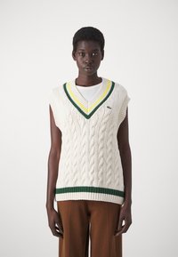 Pull sans manches en tricot crème à tresses avec un col en V rayé vert et jaune et un ourlet. Porté par-dessus un haut blanc et associé à un pantalon large marron.
