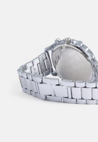 Ασημί ρολόι από ανοξείδωτο ατσάλι με σχέδιο βραχιολιού, στρογγυλή θήκη, ορατός κορώνα και κουμπιά, και ομαλή, ανακλαστική επιφάνεια.