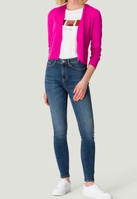 Różowy kardigan nałożony na biały t-shirt z grafiką, w zestawieniu z ciemnoniebieskimi skinny jeansami i białymi sneakersami. Gładka tekstura i dopasowany krój.