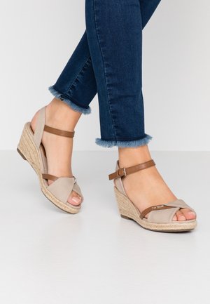 Fødder iført beige kilehælssandaler med brune stropper kombineret med mørkeblå jeans med frynset kant på hvid baggrund.