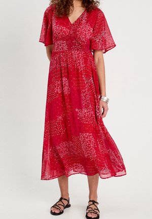 Femme portant une robe midi rouge fluide avec un motif floral subtil, des manches volantées, des sandales noires à lanières et un bracelet en argent.