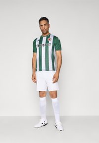 Mann som står i en grønn og hvit stripete fotballtrøye, hvite shorts, lange hvite sokker og hvite fotballsko mot en ensfarget bakgrunn.