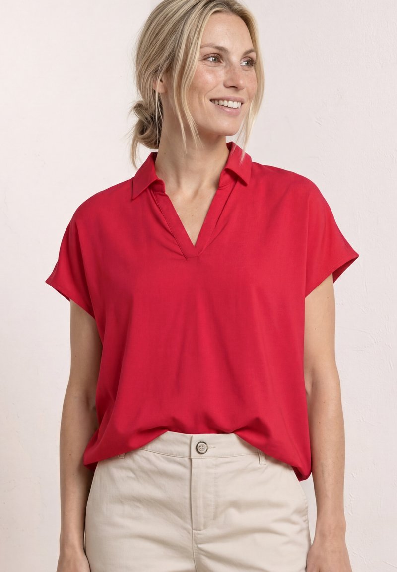 Femme blonde souriante, portant un chemisier rouge ample à manches courtes avec un col en V et un pantalon taille haute beige clair.