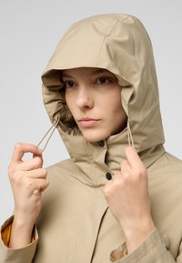 Veste imperméable beige avec capuche, dotée de cordons de serrage, de boutons-pression et d'une poche. Texture lisse avec une couleur intérieure contrastante.