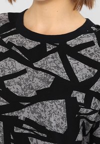 Černý a šedý grafický svetr s abstraktními, překrývajícími se vzory. Měkký materiál s kulatým výstřihem a jemným texturovaným detailem.