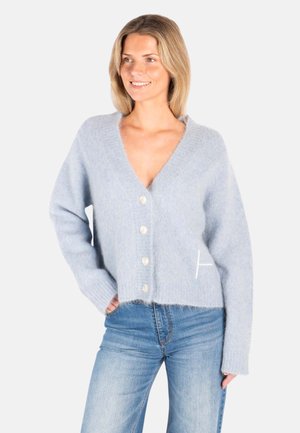 Smilende kvinne iført en lys blå, myk knappet cardigan og lys blå jeans, stående med den ene hånden i lommen mot hvit bakgrunn.