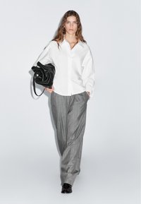 Weißes Hemd mit Knöpfen, kombiniert mit grauer Hose mit Nadelstreifen. Schwarze Handtasche mit glatter Textur, am Arm getragen. Dunkle Schuhe runden das Outfit ab.