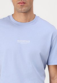 Šviesiai mėlynas medvilninis marškinėliai su apvaliu kaklu. Siūlo baltą spausdintą logotipą krūtinėje ir trumpas rankoves su lygia tekstūra.