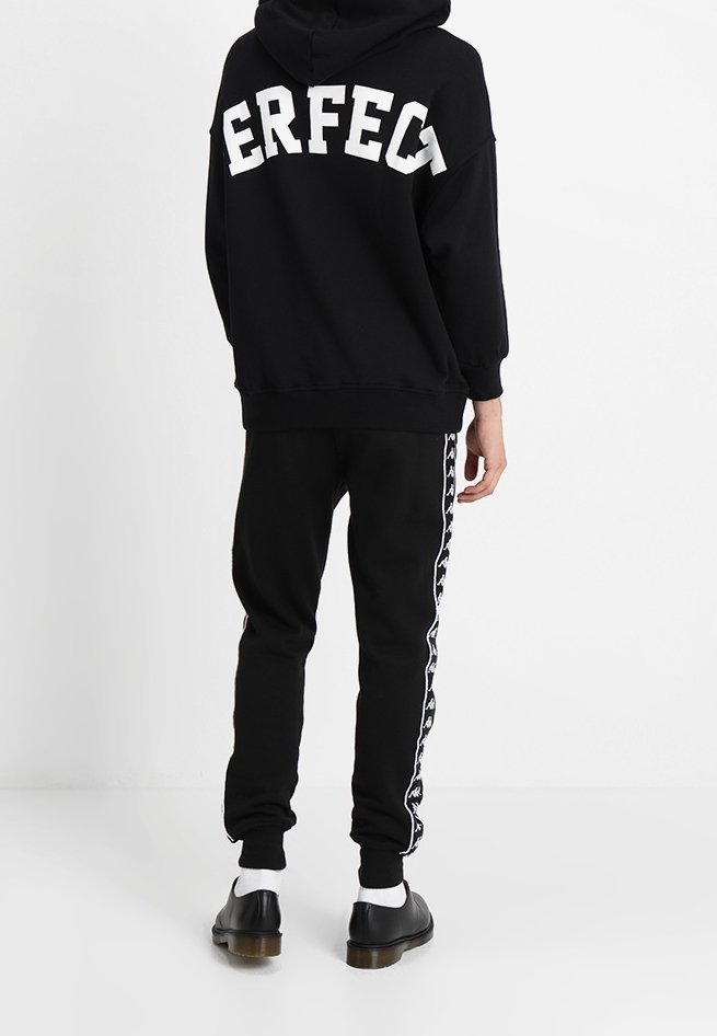 Personne portant un sweat à capuche noir avec le texte "PERFECT" au dos, un pantalon de survêtement noir à rayures blanches sur les côtés, des chaussettes blanches et des chaussures noires, debout de dos.