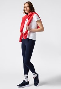 Biała koszulka z logo, granatowe legginsy, czerwony sweter zarzucony na ramiona oraz czarne buty sportowe z białą podeszwą i paskowanymi skarpetkami.