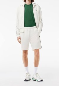 Survêtement blanc de sport avec un t-shirt vert, comprenant une veste zippée et un short, assortis à des baskets blanches et des chaussettes côtelées.