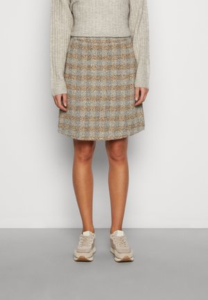 Osoba ubrana w beżową bluzkę z długim rękawem o prążkowanej fakturze, brązowo-szarą spódnicę typu A w kratę oraz beżowe trampki, stojąca na tle jednolitego jasnego tła.