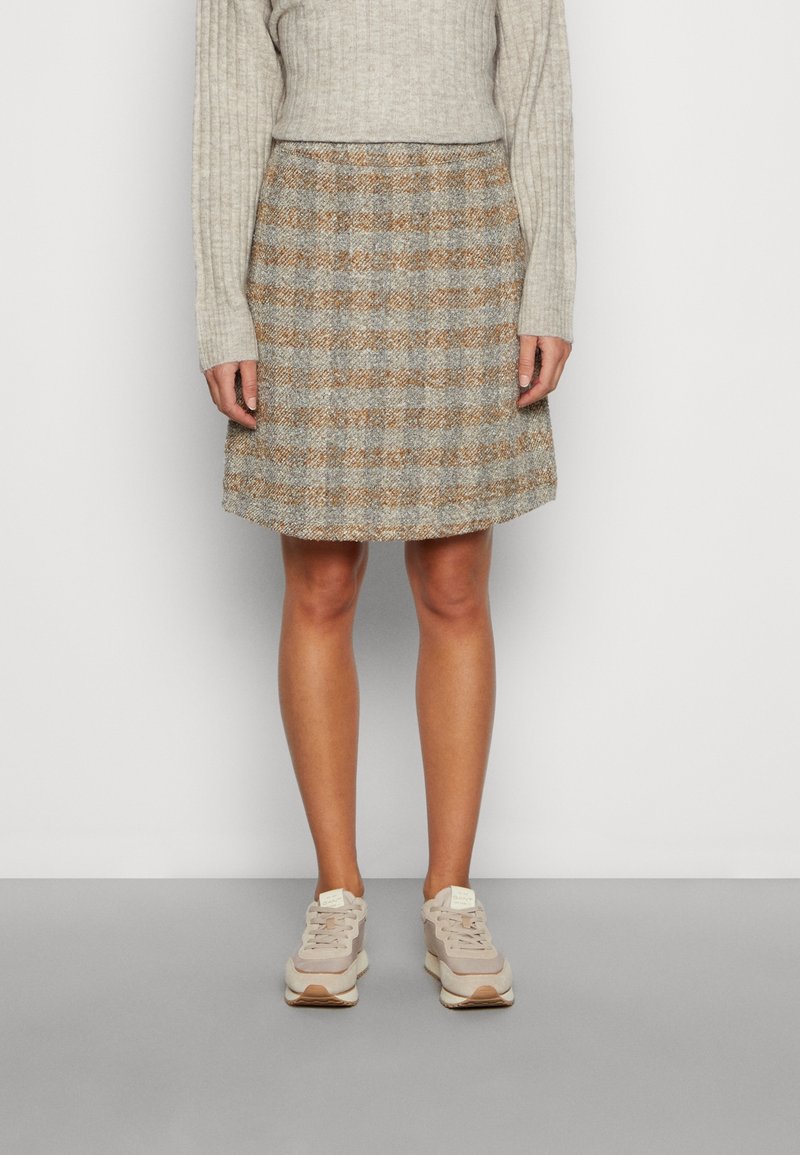 Personne portant un haut à manches longues côtelé beige, une jupe patineuse à carreaux marron et gris, et des baskets beiges, debout devant un fond clair uni.