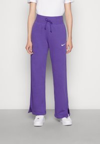 Lila, högmidjade sweatpants med dragsko, sidöppningar vid fållarna och en vit Nike-logotyp på vänster lår. Mjuk tygtextur.
