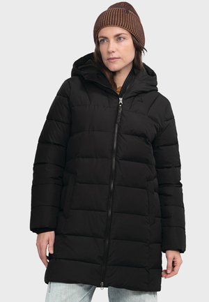 Frau trägt eine schwarze gesteppte Winterjacke mit Kapuze und eine braune gestrickte Mütze, steht mit entspannten Händen an den Seiten vor einem einfarbigen Hintergrund.