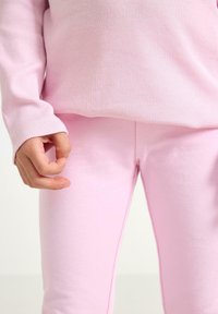 Lyserød ribbet langærmet top kombineret med matchende lyserøde leggings, med en blød tekstur og tætsiddende design. Hænderne er foldet sammen nær taljen.