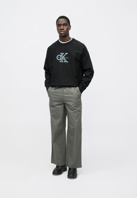 Μαύρο φούτερ με λογότυπο "CKlein", γκρι παντελόνι με φαρδιά μπατζάκια και μαύρα παπούτσια. Ομαλή υφή, casual εφαρμογή, μινιμαλιστικός σχεδιασμός.