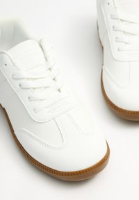 Zapatillas sintéticas blancas con una textura suave, que presentan un diseño con cordones, suela de goma marrón contrastante y detalles minimalistas.