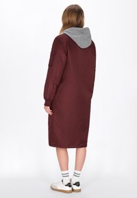 Chaqueta de plumas larga burdeos con capucha gris, que presenta un cierre frontal con cremallera, bolsillos laterales y puños elásticos. La tela tiene una textura suave.