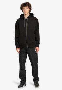 Black zip-up hoodie with a large hood, drawstrings, and front pockets. Paired with dark jeans and black athletic shoes. Casual fit.