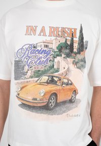 Gul vintage sportsbil med fører på en kystvei, omgitt av trær og bygninger, tekst "In a Rush Racing Club Berlin" på hvit skjorte.