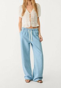 Haut blanc en dentelle à manches courtes avec un col en V et un lien à nouer à l'avant, assorti d'un pantalon bleu clair à jambes larges et cordon de serrage, et de sandales marron.