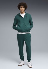 Junger Mann steht mit den Händen in den Taschen, trägt einen dunkelgrünen Kapuzenpullover und passende Jogginghosen, darunter ein weißes Shirt, sowie weiße Sneakers.