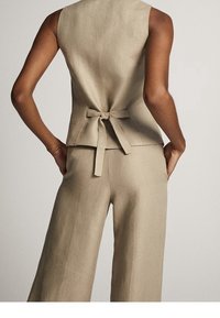 Persona vestita con un top beige senza maniche legato dietro e pantaloni ampi coordinati, in piedi con le mani nelle tasche.