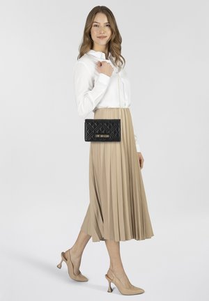Borsa a tracolla nera trapuntata con hardware dorato, tenuta da una persona che indossa una camicia bianca e una gonna plissettata beige, abbinata a décolleté nude.