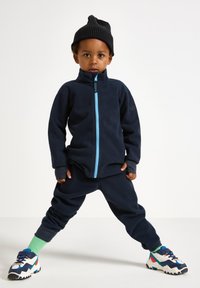 Giacca in pile navy con zip celeste, abbinata a pantaloni coordinati. Berretto lavorato a maglia nero e sneaker colorate con accenti verdi.