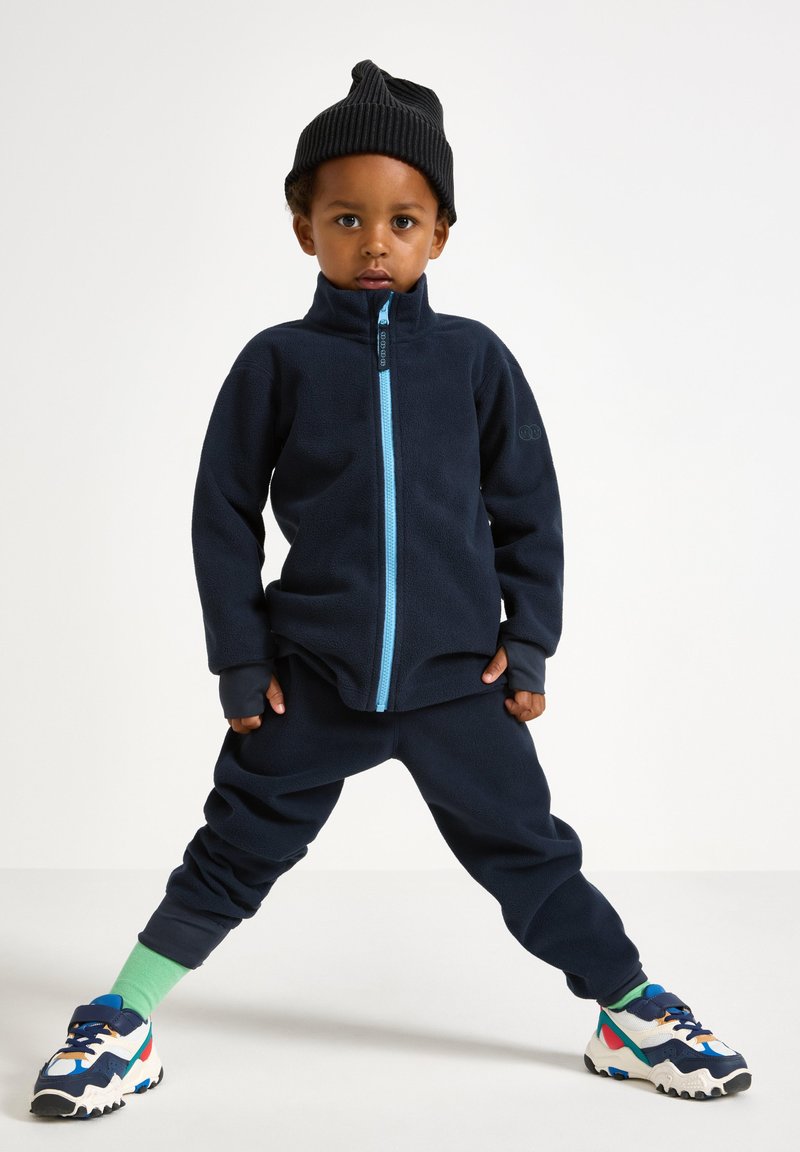 Giacca in pile navy con zip celeste, abbinata a pantaloni coordinati. Berretto lavorato a maglia nero e sneaker colorate con accenti verdi.