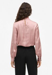 Blusa sedosa en rosa claro con un cuello alto y detalle de ojal en la parte posterior, combinada con pantalones negros ajustados y puños abotonados.