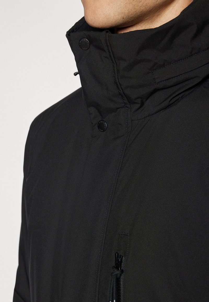 Czarna ocieplana kurtka z wysokim kołnierzem, z zapięciami na napy, kieszenią na zamek błyskawiczny po boku oraz gładkim, teksturowanym materiałem.